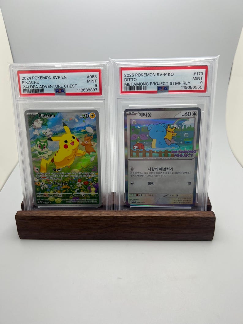 May include: Two Pokemon trading cards in protective cases, displayed on a dark wooden stand. The cards feature Pikachu and Ditto, with text including "2024 Pokemon SVP EN" and "2025 Pokemon SV-P KO". The Pikachu card has a value of 70.