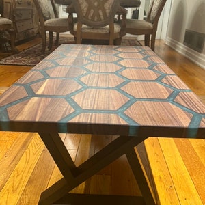 May include: A rectangular coffee table with a dark wood surface inlaid with a honeycomb pattern of teal resin. The table has a black metal base with a geometric design. The table is in a room with hardwood floors.
