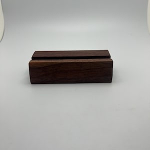 May include: A dark brown wooden phone stand with a rectangular base and a slot to hold a phone. The stand is made of solid wood and has a smooth finish. The stand is approximately 4 inches long and 1 inch tall.