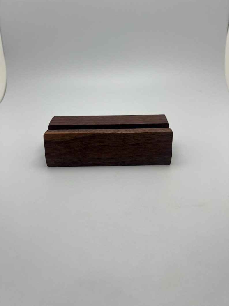 May include: A dark brown wooden phone stand with a rectangular base and a slot to hold a phone. The stand is made of solid wood and has a smooth finish. The stand is approximately 4 inches long and 1 inch tall.