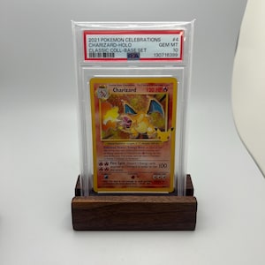May include: A graded Charizard-Holo trading card from the 2021 Pok&eacute;mon Celebrations Classic Coll-Base Set. The card is encased in a protective holder and displayed on a wooden stand. The card features a fiery orange and yellow design with the text "Charizard" and "120 HP".