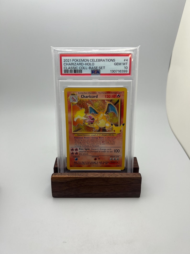 May include: A graded Charizard-Holo trading card from the 2021 Pok&eacute;mon Celebrations Classic Coll-Base Set. The card is encased in a protective holder and displayed on a wooden stand. The card features a fiery orange and yellow design with the text "Charizard" and "120 HP".