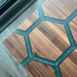 May include: Close-up of a wooden table with a geometric design. The table's surface is a rich brown, with teal-colored resin filling the hexagonal pattern. The wood grain is visible, adding texture to the design.