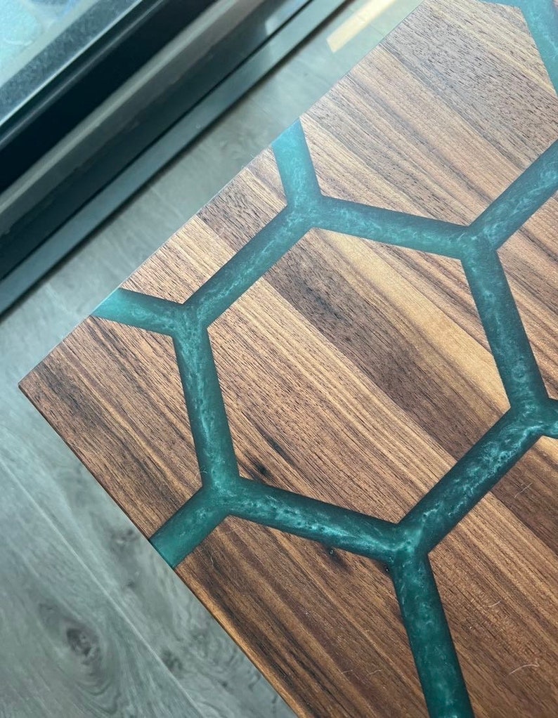 May include: Close-up of a wooden table with a geometric design. The table's surface is a rich brown, with teal-colored resin filling the hexagonal pattern. The wood grain is visible, adding texture to the design.