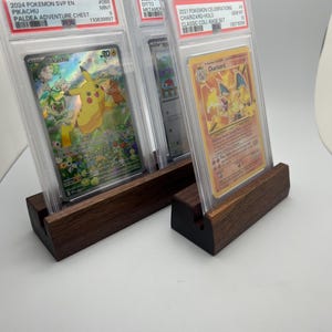 May include: Three graded Pok&eacute;mon cards in protective cases, displayed on dark wooden stands. The cards feature Pikachu and Charizard, with text including "2024 Pokemon SVP EN Pikachu" and "2021 Pokemon Celebrations Charizard-Holo".