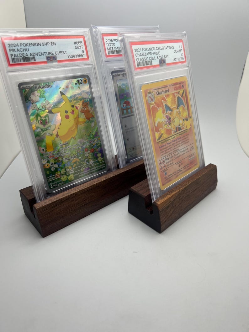 May include: Three graded Pok&eacute;mon cards in protective cases, displayed on dark wooden stands. The cards feature Pikachu and Charizard, with text including "2024 Pokemon SVP EN Pikachu" and "2021 Pokemon Celebrations Charizard-Holo".