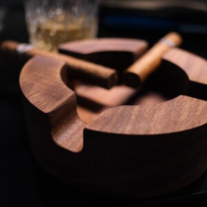 May include: A dark brown wooden cigar ashtray with two cigars resting inside. The ashtray has a rounded shape and a smooth finish.