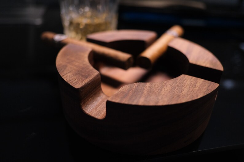 May include: A dark brown wooden cigar ashtray with two cigars resting inside. The ashtray has a rounded shape and a smooth finish.