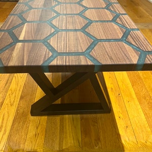 May include: A rectangular wooden coffee table with a unique honeycomb design. The tabletop features dark wood sections separated by teal-colored resin. The table has a black metal base with a geometric design, and is set on a hardwood floor.