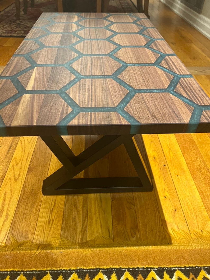 May include: A rectangular wooden coffee table with a unique honeycomb design. The tabletop features dark wood sections separated by teal-colored resin. The table has a black metal base with a geometric design, and is set on a hardwood floor.