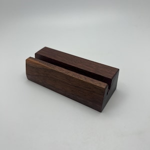 May include: A dark brown wooden phone stand with a rectangular base and a slot to hold a phone. The stand is made of solid wood and has a natural finish. The stand is designed to hold a phone in a vertical position.