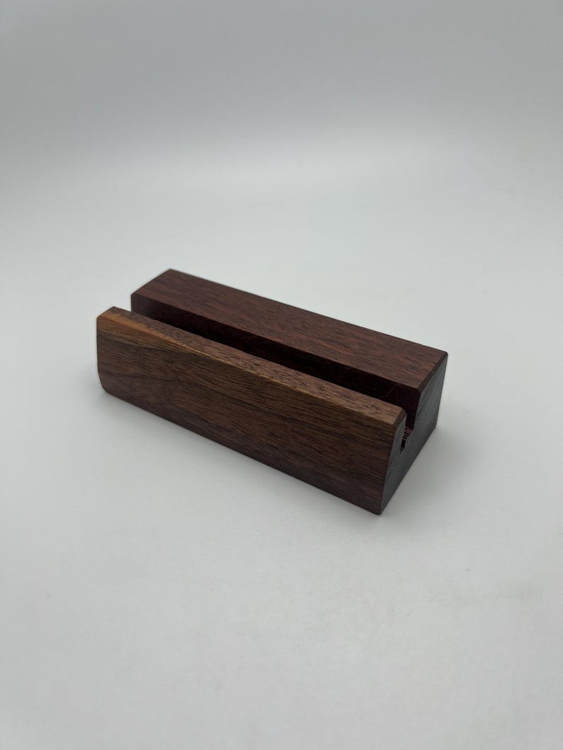 May include: A dark brown wooden phone stand with a rectangular base and a slot to hold a phone. The stand is made of solid wood and has a natural finish. The stand is designed to hold a phone in a vertical position.