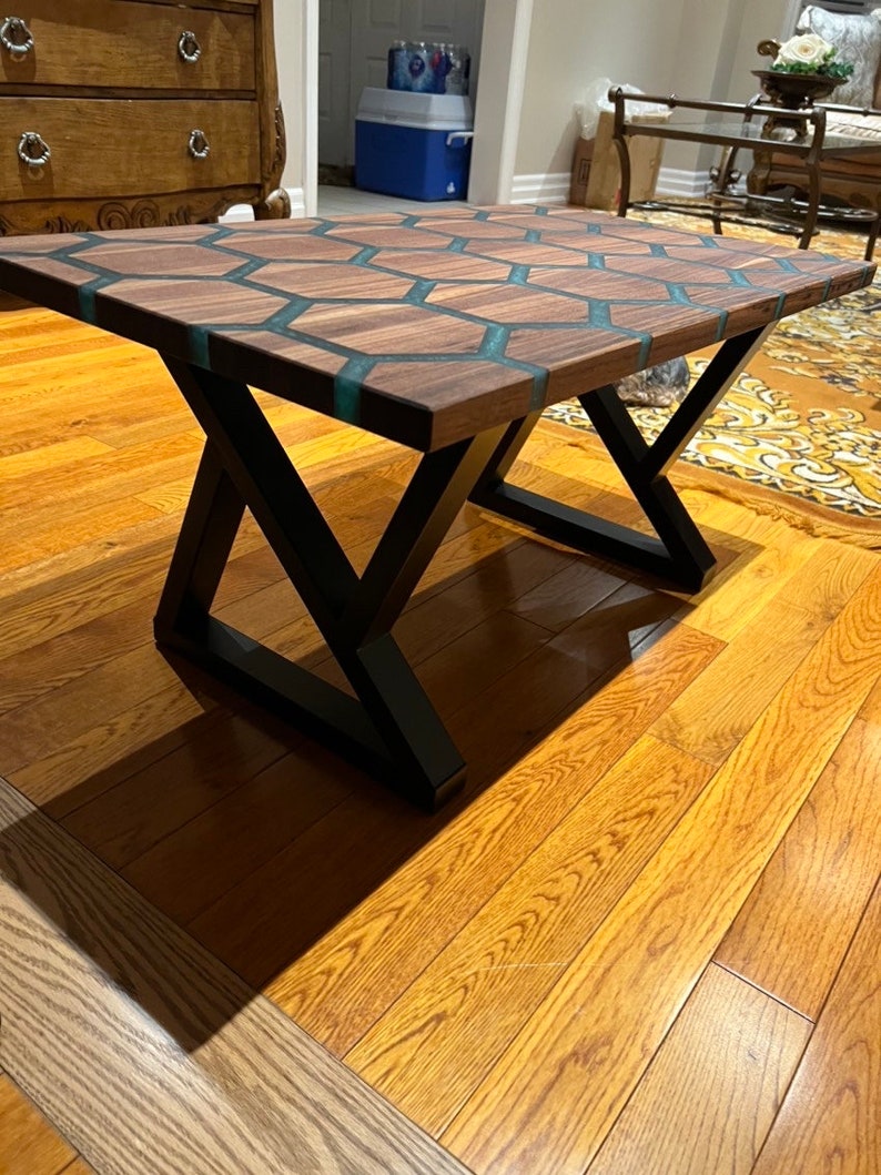 May include: A rectangular coffee table with a dark wood surface featuring a honeycomb design with teal resin inlays. The table has black metal X-shaped legs and sits on a hardwood floor.