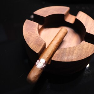 May include: A wooden cigar ashtray with a brown cigar resting inside. The ashtray has a circular shape with a deep well in the center.