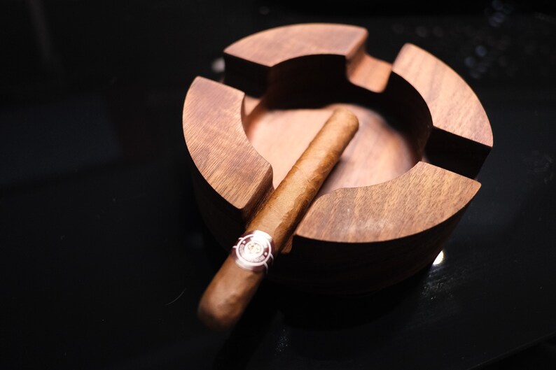 May include: A wooden cigar ashtray with a brown cigar resting inside. The ashtray has a circular shape with a deep well in the center.