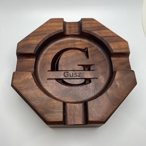 May include: A wooden ashtray with a dark brown finish. The ashtray has an octagonal shape with a circular indentation in the center. The letter "G" and the name "Gusz" are engraved in the center of the ashtray.