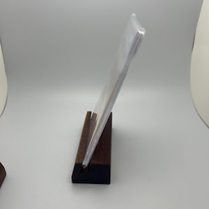 May include: A clear acrylic display stand holding a clear plastic sleeve. The stand is made of dark brown wood with a black base. The display is angled, showcasing the contents within the sleeve.