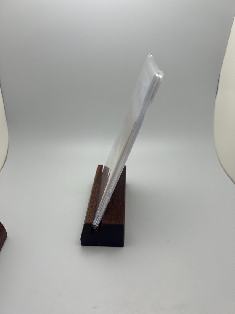 May include: A clear acrylic display stand holding a clear plastic sleeve. The stand is made of dark brown wood with a black base. The display is angled, showcasing the contents within the sleeve.