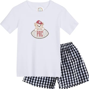 May include: White short-sleeved t-shirt and black and white checkered shorts. The shirt features an embroidered design of a bulldog wearing a maroon hat and holding a football with the letters "PRC". The shorts have an elastic waistband.
