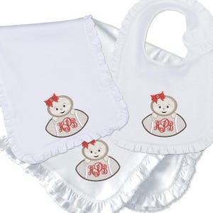 Könnte beinhalten: Weißes Baby-Set mit Decke, Lätzchen und Spucktuch, alle mit Rüschenkanten. Jedes Teil hat ein gesticktes Puppen-Design mit roter Schleife und rot monogrammierten Initialen.