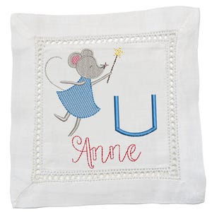 May include: White square linen coaster with a whimsical embroidered design. It features a gray mouse in a blue dress holding a golden wand, the name "Anne" in red script, and a blue letter "U". The coaster has a delicate lace border.