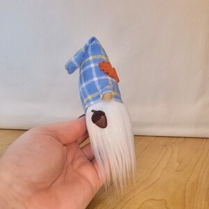 May include: A handmade gnome doll with a white beard and a blue plaid hat. The hat is adorned with an orange leaf and the gnome has a brown acorn-shaped nose. The doll is a decorative item.