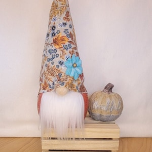 May include: A decorative gnome with a long white beard and a floral patterned hat. A blue flower adorns the hat. Beside the gnome is a small pumpkin with the word "thankful" on it. The gnome and pumpkin sit on a wooden crate.