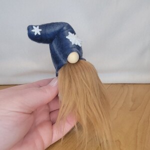 May include: A handmade gnome doll with a navy blue hat adorned with white snowflake accents. The gnome has a light brown, fluffy beard and a wooden nose. The doll is held in a hand.