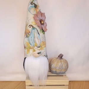 May include: A decorative gnome with a long white beard and a tall, patterned hat featuring pumpkins and fall foliage. A small pumpkin with the word "thankful" sits beside the gnome on a wooden crate. The gnome has a wooden nose and a flower.