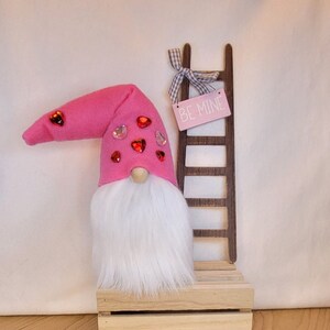 May include: A pink gnome with a white beard and red heart embellishments sits on a wooden crate with a wooden ladder behind it. The ladder has a pink sign that says "Be Mine".