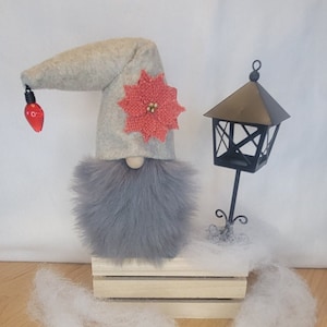 May include: A whimsical Christmas gnome with a gray felt hat adorned with a red poinsettia and a dangling red lightbulb. The gnome has a fluffy gray beard and sits on a wooden crate next to a black metal lantern.