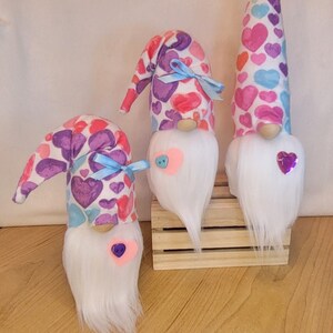 May include: Three Valentine's Day gnomes with white beards and heart-shaped accents. The gnomes have patterned hats with pink, purple, and blue hearts. One gnome has a tall, pointed hat, while the others have curved hats. Each gnome has a wooden nose and a heart-shaped embellishment.