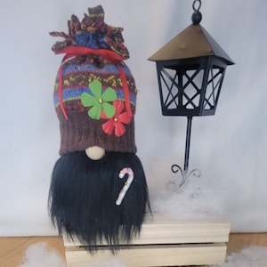May include: A handmade Christmas gnome with a black beard, a patterned hat, and a candy cane accent. A black lantern stands beside the gnome on a wooden crate, with decorative snow around the base. Festive holiday decor.