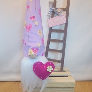 May include: A Valentine's Day gnome with a lavender hat patterned with pink and yellow trees and flowers. The gnome has a white beard and a pink felt heart with a flower button. A small wooden ladder with a sign that says "BE MINE" is behind the gnome.