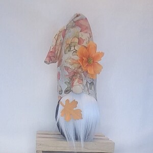 May include: A fall-themed gnome figurine with a tall, patterned hat adorned with orange leaves and a flower. The gnome has a white beard, a wooden nose, and an orange leaf accent. It sits on a small wooden crate.