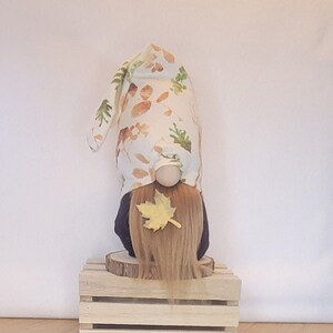 May include: A fall-themed gnome figurine with a long, light brown beard and a yellow leaf accent. The gnome wears a white hat patterned with brown and green leaves. It stands on a small wooden crate and a wood slice base.