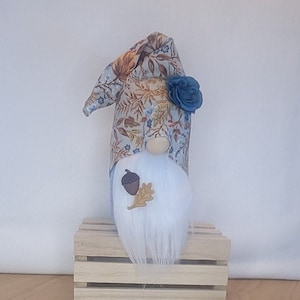 May include: A white-bearded gnome with a blue and brown floral hat, a blue flower, and an acorn and leaf accent. The gnome is sitting on a wooden crate.