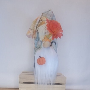 May include: A fall-themed gnome doll with a white beard and a small orange pumpkin attached to the front. The gnome wears a patterned hat with fall leaves and a large orange flower. It sits on a small wooden crate.