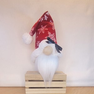 May include: A Christmas gnome doll with a red hat patterned with white snowflakes and a white fluffy beard. The gnome has a wooden nose and a black bow. It is sitting on a small wooden crate.