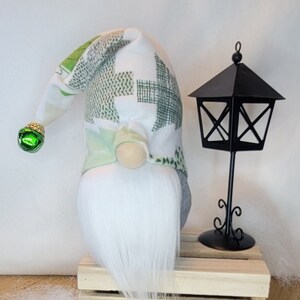 May include: A handmade gnome with a white faux fur beard and a patchwork hat in shades of green and white. The gnome has a small, round nose and a green bell on the end of its hat. A black metal lantern sits beside the gnome on a wooden crate.