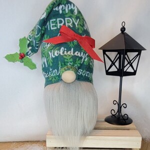 May include: A Christmas gnome with a long gray beard and a green hat adorned with the words "Happy Merry Christmas Holidays Seasons". The hat is decorated with white snowflakes and a red bow. A black lantern sits next to the gnome.