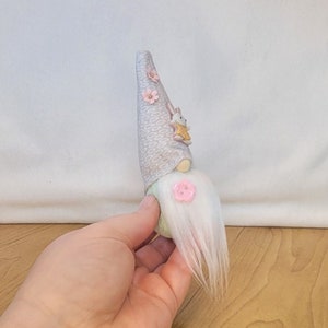May include: A light green and gray gnome with a white beard and pink flowers on its hat. A small bunny is attached to the gnome's hat.