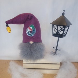 May include: A Christmas gnome with a burgundy hat, gray beard, and a decorative wreath. The hat has a yellow light bulb attached. A black metal lantern sits nearby on a wooden crate, with faux snow around it.
