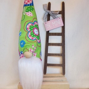 May include: A Valentine's Day gnome with a long white beard, a wooden nose, and a green hat patterned with flowers and buttons. A small wooden ladder with a pink sign that says "BE MINE" is next to the gnome.