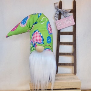 May include: A Valentine's Day gnome with a long white beard and a green hat patterned with hearts and gingham. The gnome sits on a wooden crate next to a miniature ladder with a "BE MINE" sign.