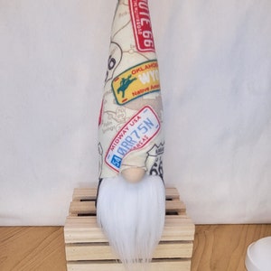 May include: A white gnome with a long white beard wearing a hat with a Route 66 theme. The hat is covered in patches with various Route 66 logos and text, including "Route 66", "Midway USA", "OKLAHOMA", "WYO", "Native American", and "54 ARR 75N KANSAS".
