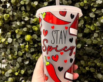 Op de afbeelding: Een witte herbruikbare beker met een grijze achtergrond en rode pepers en zwarte harten. De beker heeft de tekst "Stay Spicy" in zwarte letters.