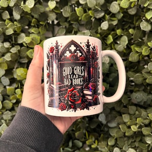 May include: White ceramic mug with a gothic-style design featuring a black archway with red roses and the text "GOOD GIRLS READ BAD BOOKS".