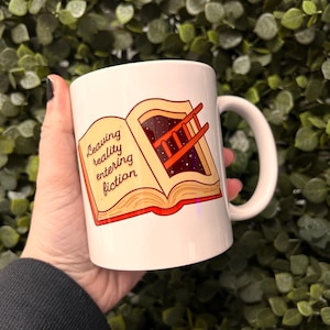 May include: White ceramic mug featuring a colourful illustration of an open book with a ladder leading into a starry sky. The text on the mug reads "Leaving reality entering fiction."