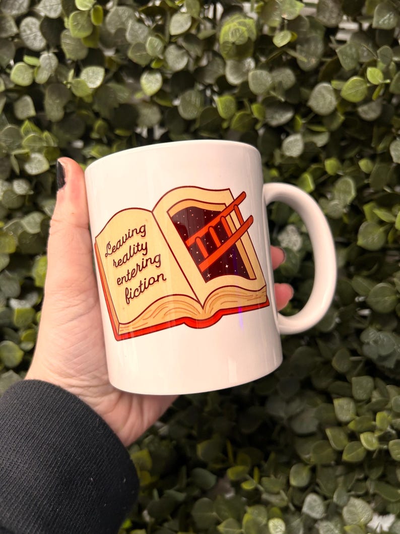 May include: White ceramic mug featuring a colourful illustration of an open book with a ladder leading into a starry sky. The text on the mug reads "Leaving reality entering fiction."